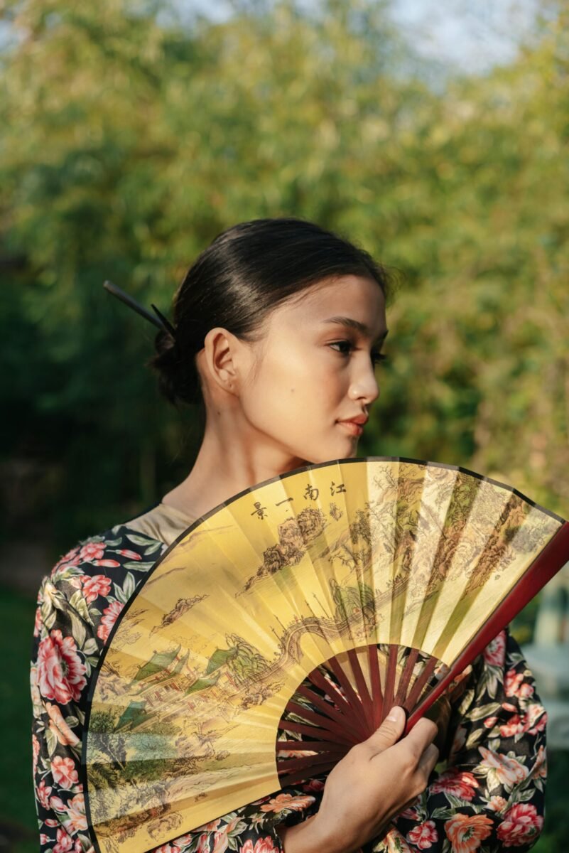 Charming woman holding a floral fan outdoors, exuding elegance and grace.