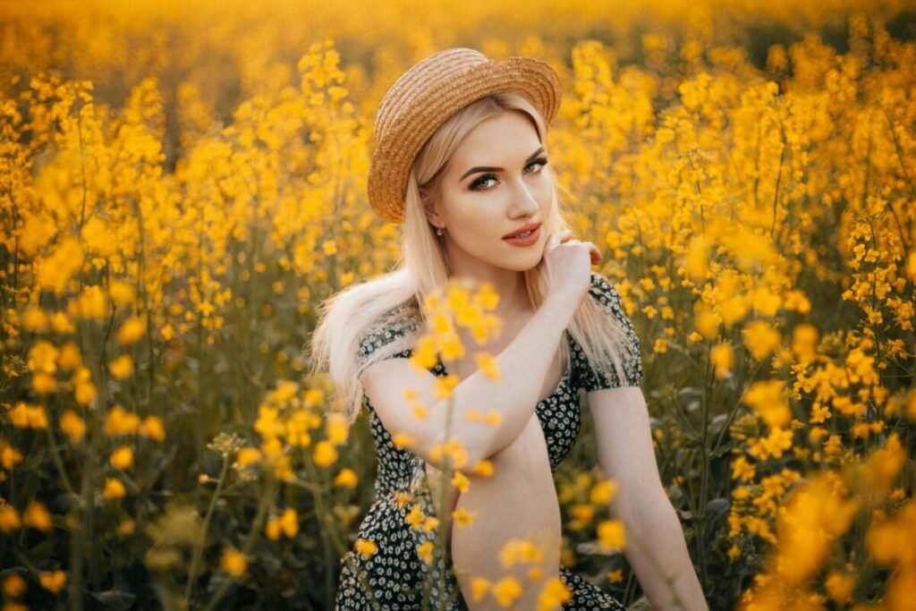 Blonde woman in floral dress sitting in a field of yellow flowers, wearing a straw hat.